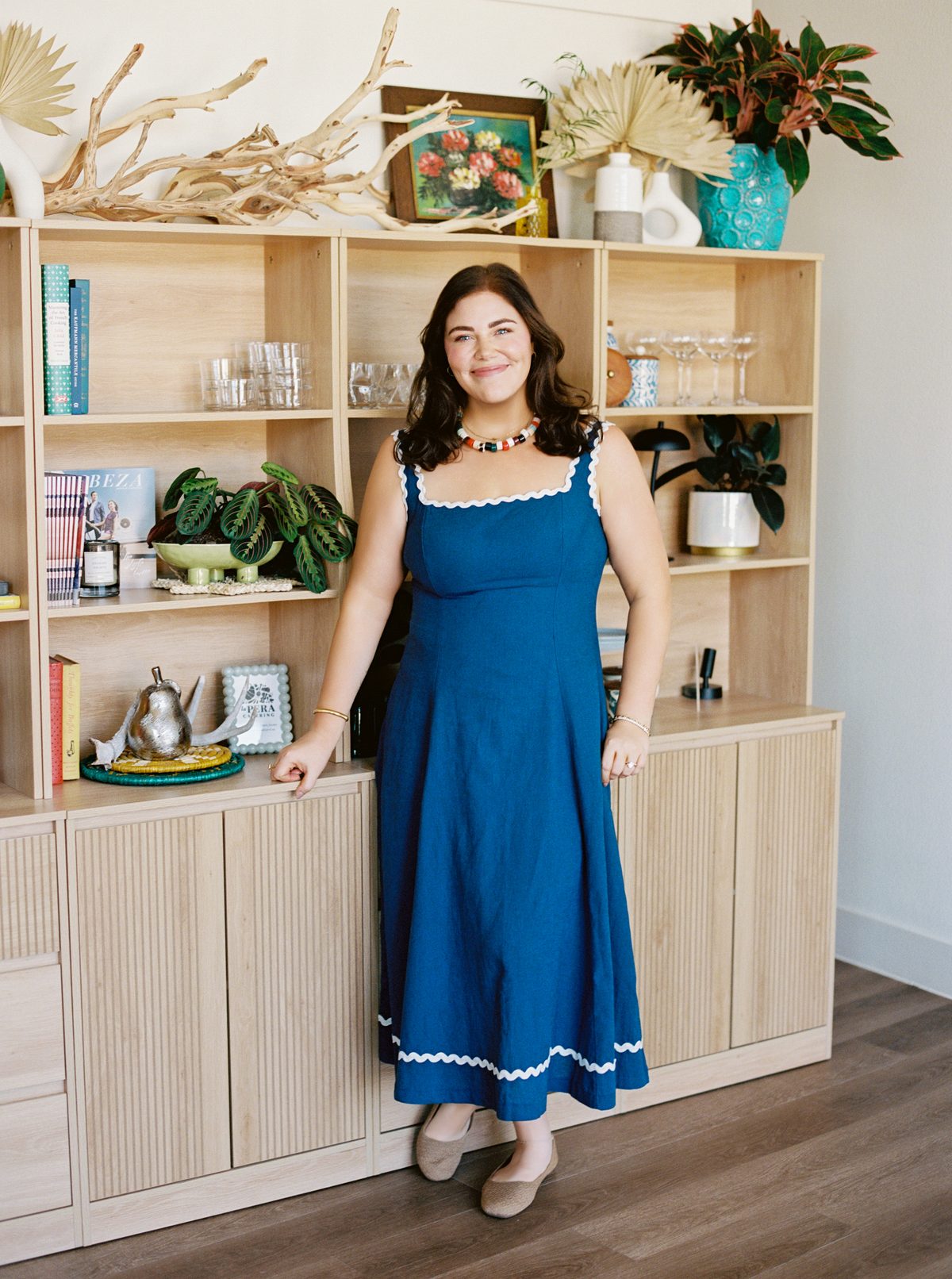 Paige Vaughn standing and smiling in front of a light-wood shelf styled with plants, glassware, and ceramics
